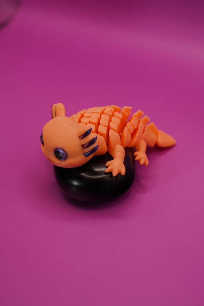 3D printed orange Flexolotl (flexible articulated axolotl figurine) displayed on a black round base